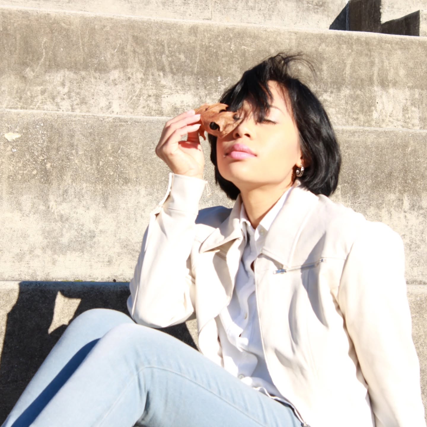 Dorothy Tobias-Williams sitting on concrete steps in a cream jacket and light jeans, holding sunglasses near her face.