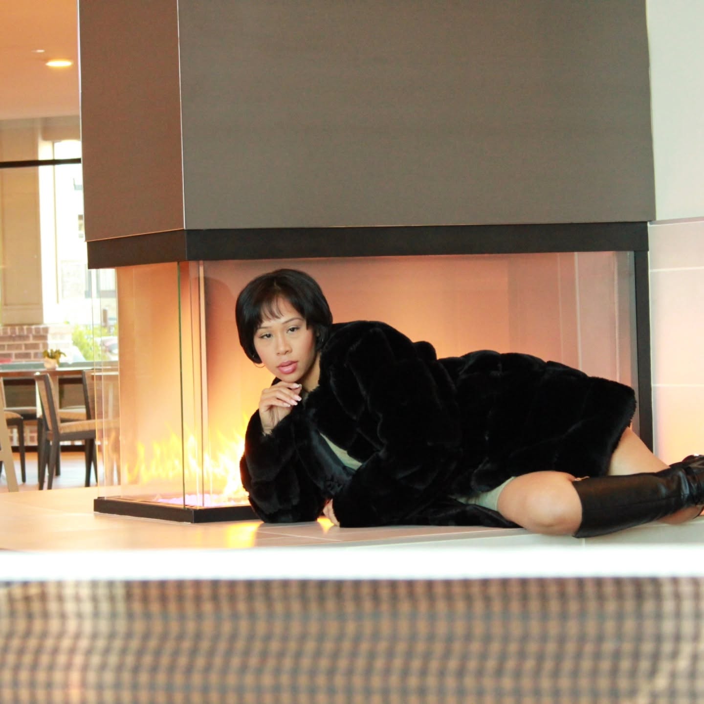 Dorothy Tobias-Williams posing in a black faux-fur coat and tall boots beside a modern indoor fireplace.